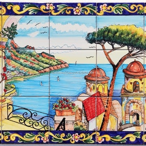 May include: A colorful hand-painted ceramic tile mural depicting a coastal Italian scene. The mural features a view of a bay with sailboats, a town with buildings, and a lush green hillside. The mural is framed with a blue and gold border with floral accents.