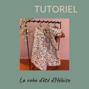 Puede incluir: Un vestido de estampado floral y un cárdigan en un perchero de metal negro. El vestido tiene un fondo blanco con flores rojas, naranjas y amarillas. El cárdigan es de color rosa sólido. El texto "La robe d'été d'Héloïse" está debajo del perchero.