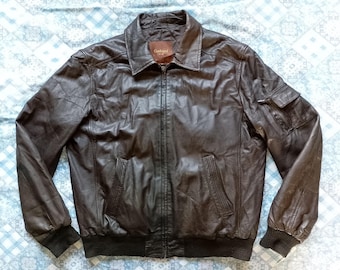 Puede incluir: Chaqueta de cuero marrón con cuello, cierre de cremallera y múltiples bolsillos. La chaqueta tiene puños y dobladillo acanalados. El cuero parece desgastado, dándole un aspecto vintage. La chaqueta es de color marrón medio.