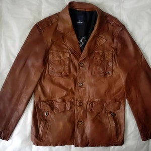 Puede incluir: Una chaqueta de cuero marrón con cierre de botones, múltiples bolsillos y un detalle con cremallera en la parte inferior.