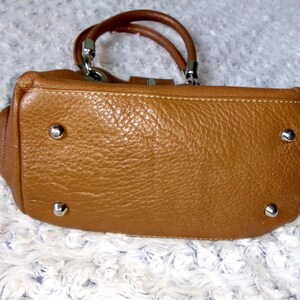 May include: A tan leather handbag with a textured surface. The bag has two brown handles and silver-colored hardware. The bottom of the bag has four silver-colored metal feet.