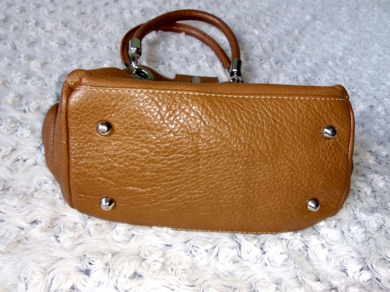 May include: A tan leather handbag with a textured surface. The bag has two brown handles and silver-colored hardware. The bottom of the bag has four silver-colored metal feet.