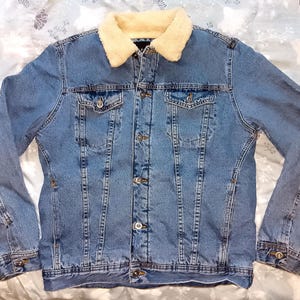 May include: A light blue denim jacket with a cream-coloured, faux-shearling collar. The jacket has two chest pockets, two side pockets, and a button-up front. The sleeves have buttoned cuffs. A classic denim jacket.