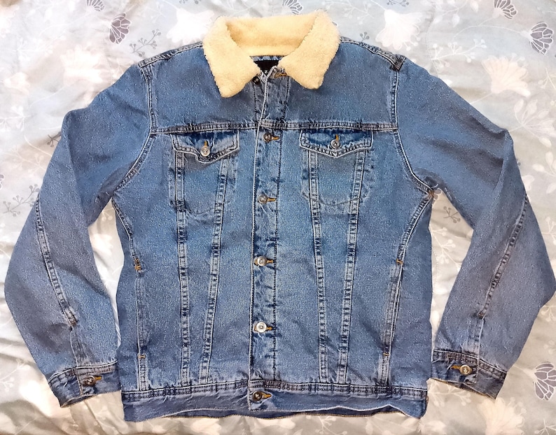 May include: A light blue denim jacket with a cream-coloured, faux-shearling collar. The jacket has two chest pockets, two side pockets, and a button-up front. The sleeves have buttoned cuffs. A classic denim jacket.