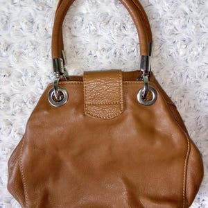 May include: A brown leather handbag with a rounded shape and a top handle. The bag features silver-tone hardware, including rings connecting the handle to the body. The bag has a textured flap closure.