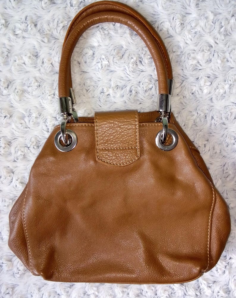 May include: A brown leather handbag with a rounded shape and a top handle. The bag features silver-tone hardware, including rings connecting the handle to the body. The bag has a textured flap closure.