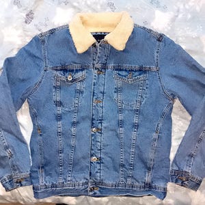 May include: A light blue denim jacket with a cream-coloured, fluffy collar. The jacket features two chest pockets, button fastenings, and a classic design. This outerwear is suitable for casual wear.