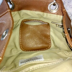 May include: Brown leather handbag with a light tan interior. The bag features a small, stitched pocket and a zipper closure. Silver-tone hardware includes rings and a clasp. The bag is made of genuine leather.