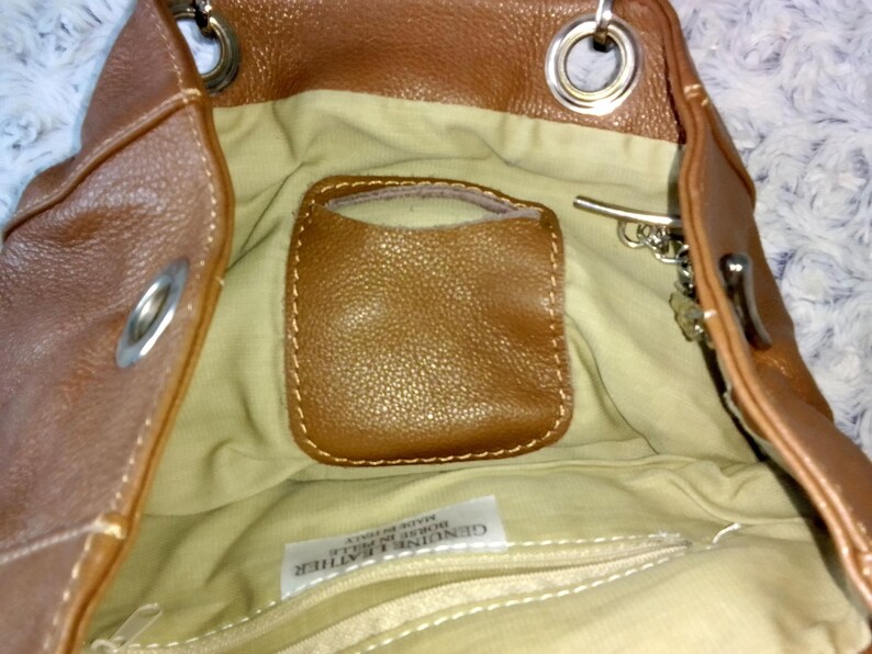 May include: Brown leather handbag with a light tan interior. The bag features a small, stitched pocket and a zipper closure. Silver-tone hardware includes rings and a clasp. The bag is made of genuine leather.