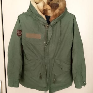 Może przedstawiać: Oliwkowozielona parka z kapturem wyłożonym futrem. Kurtka posiada zapięcie na zamek błyskawiczny, liczne kieszenie oraz naszywkę na lewym górnym rękawie. Wnętrze kaptura jest w kolorze jasnobrązowym.