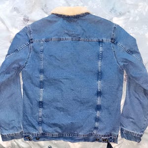 May include: A light blue denim jacket with a cream-coloured shearling collar. The jacket has a classic design with button fastenings, pockets, and a tailored fit. The back view shows the jacket's construction and details.