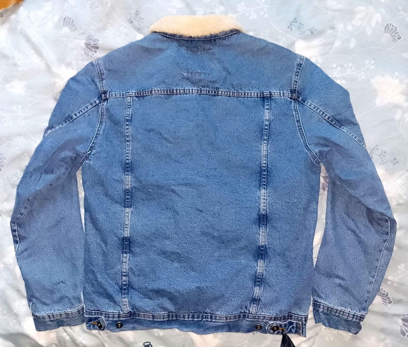 May include: A light blue denim jacket with a cream-coloured shearling collar. The jacket has a classic design with button fastenings, pockets, and a tailored fit. The back view shows the jacket's construction and details.