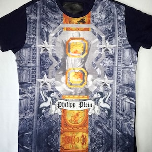 Puede incluir: Una camiseta azul marino con un estampado gráfico de una escena religiosa con detalles dorados. El estampado presenta una figura con un halo, estrellas y el texto "Philipp Plein".