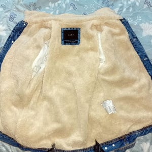 May include: A denim jacket in light blue, with a beige, fluffy interior lining. The jacket features a button-up front and a small label reading "ZARA". The jacket is open, displaying the soft lining.