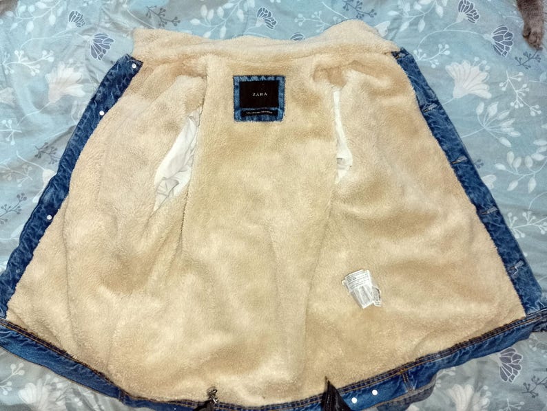 May include: A denim jacket in light blue, with a beige, fluffy interior lining. The jacket features a button-up front and a small label reading "ZARA". The jacket is open, displaying the soft lining.