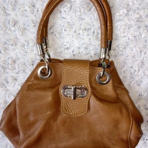 May include: A brown leather handbag with a rounded shape and two rolled handles. The bag features silver-tone hardware, including rings connecting the handles and a clasp closure on the front. The leather has a textured appearance.