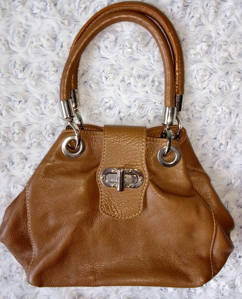 May include: A brown leather handbag with a rounded shape and two rolled handles. The bag features silver-tone hardware, including rings connecting the handles and a clasp closure on the front. The leather has a textured appearance.