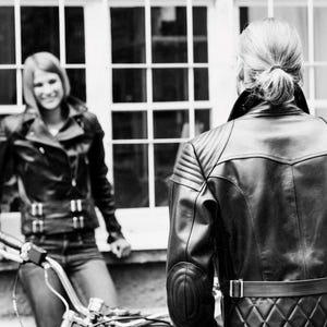 May include: Black leather motorcycle jackets with quilted and padded details. One person is leaning on a motorcycle, while another is facing away from the camera. The jackets have multiple zippers and a belt at the waist. The image is in black and white.