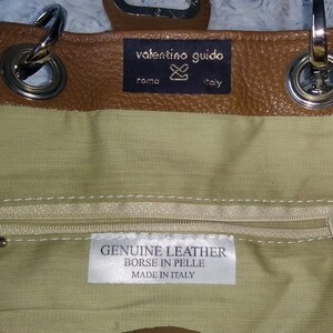 May include: A brown leather handbag with silver hardware. The interior is a light green fabric with a zipper pocket. The label reads "GENUINE LEATHER BORSE IN PELLE MADE IN ITALY". The brand name "valentino guido" is visible.