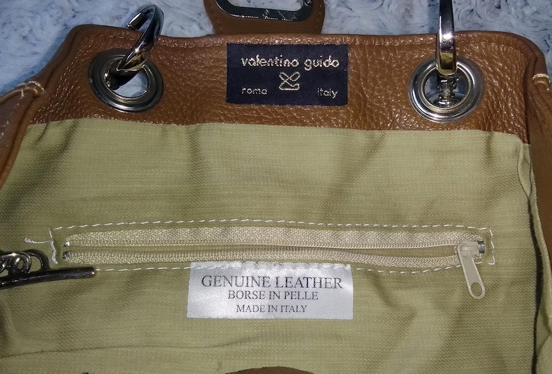 May include: A brown leather handbag with silver hardware. The interior is a light green fabric with a zipper pocket. The label reads "GENUINE LEATHER BORSE IN PELLE MADE IN ITALY". The brand name "valentino guido" is visible.