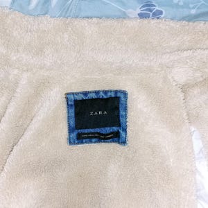 May include: Close-up of a denim label with the brand name "ZARA" on a cream-coloured, fuzzy fabric. The label is rectangular with a dark blue background and a lighter blue denim border. The fabric appears to be the lining of a jacket.
