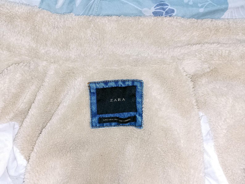 May include: Close-up of a denim label with the brand name "ZARA" on a cream-coloured, fuzzy fabric. The label is rectangular with a dark blue background and a lighter blue denim border. The fabric appears to be the lining of a jacket.