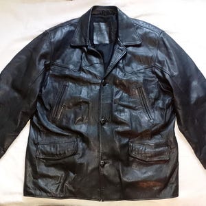 Puede incluir: Chaqueta de cuero negro con cierre de botones, dos bolsillos delanteros y cuello. La chaqueta tiene mangas largas con puños abotonados. El cuero parece ligeramente desgastado, dándole un aspecto vintage. La chaqueta está tendida sobre una superficie blanca.