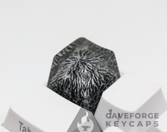 May include: A black and white keycap with a unique, swirling pattern. The keycap is made of clear resin and has a glossy finish. The keycap is sitting on a white keyboard with the word "Tab" visible.