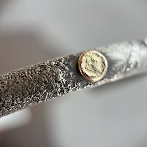 May include: A silver bracelet with a textured surface and a round, gold-colored accent.