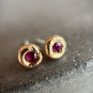 May include: A pair of gold stud earrings, each featuring a round, textured design. The center of each earring holds a vibrant, round, ruby-colored stone. The earrings are set against a neutral, textured background.