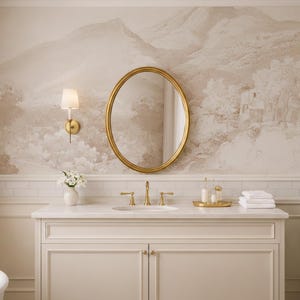 May include: A bathroom interior with a gold oval mirror, white vanity unit with gold taps, and a landscape mural. A small vase of white flowers sits on the vanity, along with toiletries and white towels. A potted plant is in the corner.