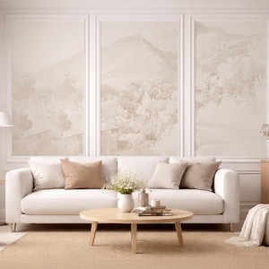 May include: A living room interior with a white sofa, beige cushions, and a round wooden coffee table. The wall features three framed landscape paintings in neutral tones. A beige rug, floor lamp, vase with pampas grass, and side table complete the scene.