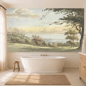 May include: A large wall mural featuring a landscape painting of a grassy hill, trees, and a calm body of water with a small boat under a soft sky. The mural is positioned behind a white freestanding bathtub in a bathroom with light wood cabinetry.