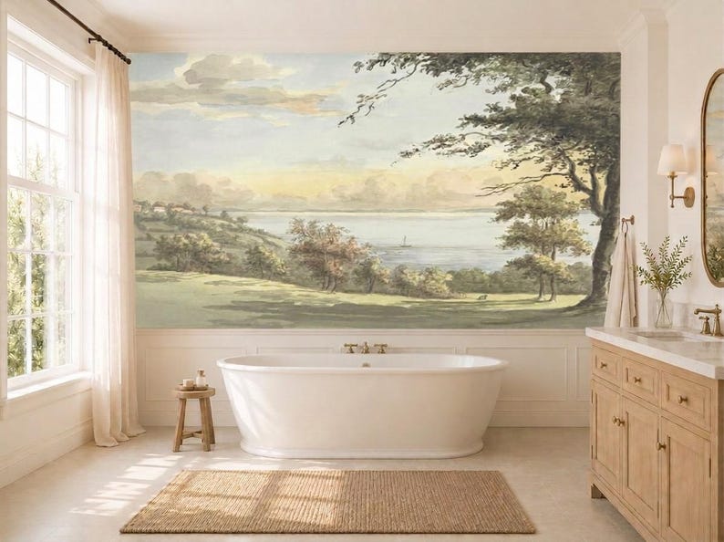 May include: A large wall mural featuring a landscape painting of a grassy hill, trees, and a calm body of water with a small boat under a soft sky. The mural is positioned behind a white freestanding bathtub in a bathroom with light wood cabinetry.