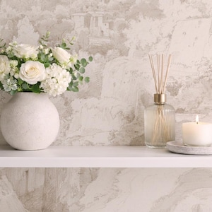 May include: A white ceramic vase filled with white roses and foliage sits on a white shelf. A glass diffuser with reeds and a lit candle on a stone plate are also on the shelf. The background is a neutral wallpaper.