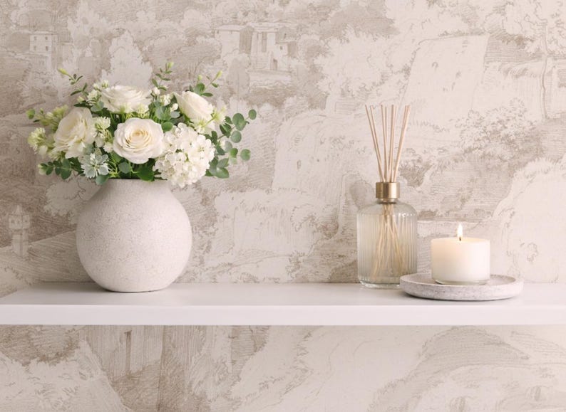 May include: A white ceramic vase filled with white roses and foliage sits on a white shelf. A glass diffuser with reeds and a lit candle on a stone plate are also on the shelf. The background is a neutral wallpaper.