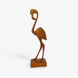 Könnte beinhalten: Eine Holzskulptur eines Flamingos, der auf einem quadratischen Sockel steht. Der Flamingo ist in einem stilisierten, abstrakten Stil geschnitzt, mit einem langen Hals und schlanken Beinen. Die Skulptur ist in einem warmen Braunton bemalt.