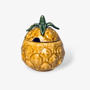 May include: A ceramic pineapple-shaped sugar bowl with a green leaf lid. The bowl is glazed in a light brown color and has a textured surface.