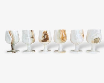 May include: Six onyx wine glasses with a white and brown marbled pattern. The glasses have a rounded bowl shape and a stem. The natural stone has a unique pattern, making each glass distinct.