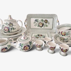 Op de afbeelding: Een keramieken theeservies met een witte basis, een veelkleurig bloemmotief en groene stippenranden. De set bevat een theepot, een suikerpot met deksel, een melkkan, twee rechthoekige dienbladen, vier eierdopjes en diverse borden en theekopjes.