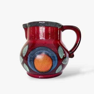 May include: A small, red ceramic jug with a rounded body and a handle. The jug features a design of a blue circle with an orange centre, surrounded by green and blue leaf-like shapes. The rim is dark grey.