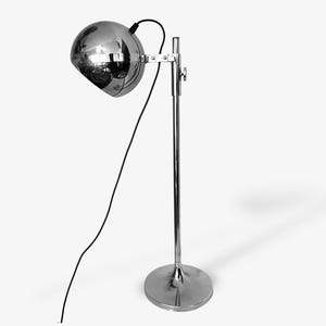 Op de afbeelding: Een verchroomde vloerlamp met een bolvormige kop. De lamp heeft een ronde voet en een slanke, verstelbare paal. Een zwart netsnoer loopt van de lampkop. De lamp is ontworpen voor gebruik binnenshuis.