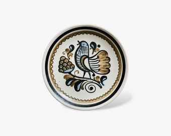 May include: A white ceramic plate with a brown and blue bird design. The bird is perched on a branch with a cluster of berries. The plate has a brown rim with a decorative pattern.