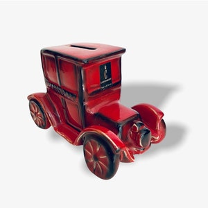 Könnte beinhalten: Rote Keramik-Sparbüchse in Form eines Oldtimers mit schwarzem und goldenem Besatz. Das Auto hat einen Schlitz auf dem Dach für Münzen und die Worte "C. CHRISTOPHER" sind an der Seite aufgedruckt.