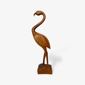 Könnte beinhalten: Eine Holzschnitzerei eines Flamingos, der auf einem Sockel steht. Der Flamingo ist braun und hat einen langen Hals und lange Beine.