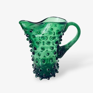 Könnte beinhalten: Eine grüne Glaskanne mit einer strukturierten, unebenen Oberfläche. Die Kanne hat eine breite Öffnung und einen Henkel.