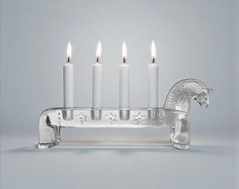 May include: A clear glass candle holder shaped like a horse, holding four white candles with lit flames. The horse design is detailed, and the holder has a decorative pattern. The background is a soft, neutral grey.