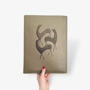 Puede incluir: Un portafolio rectangular de cuero verde oliva con un diseño en relieve de tres ciervos estilizados con contornos marrón oscuro. El portafolio es sostenido por una persona. El diseño es abstracto.