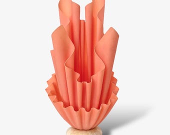 May include: A coral-colored, abstract sculpture with a ruffled, paper-like texture. The sculpture is mounted on a small, round, beige stone base. The design features vertical, folded elements that create a dynamic, artistic form.