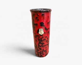 Może przedstawiać: Cylindryczny wazon ceramiczny z żywą czerwoną glazurą i nakrapianymi czarnymi wzorami na całej powierzchni. Mały, metalowy emblemat w kształcie tarczy z napisem "Scheurich" jest przymocowany na środku przedniej części wazonu.